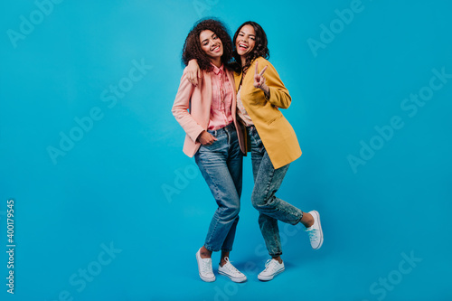 Brunette latin girl posing in studio with friend. Slim ladies in jeans standing on blue background.