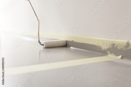 Close-up of a paint roller applying a new layer of grey paint on a floor, with the plinth protected with yellow painters tape.
