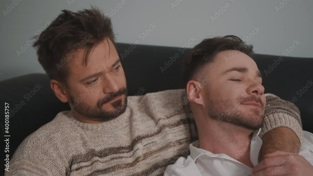 Close up. Two gay men in casual outfits sitting on sofa embracing and watching TV at home. High quality 4k footage Close up. Two gay men in casual outfits sitting on sofa embracing and watching TV at home. High quality 4k footage