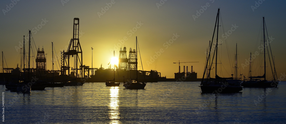 Sunrise from the bay with anchored sailboats in the foreground and commercial port in the background Sunrise from the bay with anchored sailboats in the foreground and commercial port in the background