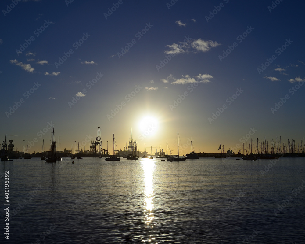 Sunrise from the bay with many sailboats and port in the background, calm sea and blue sky with small clouds, Las Palmas of Gran Canaria