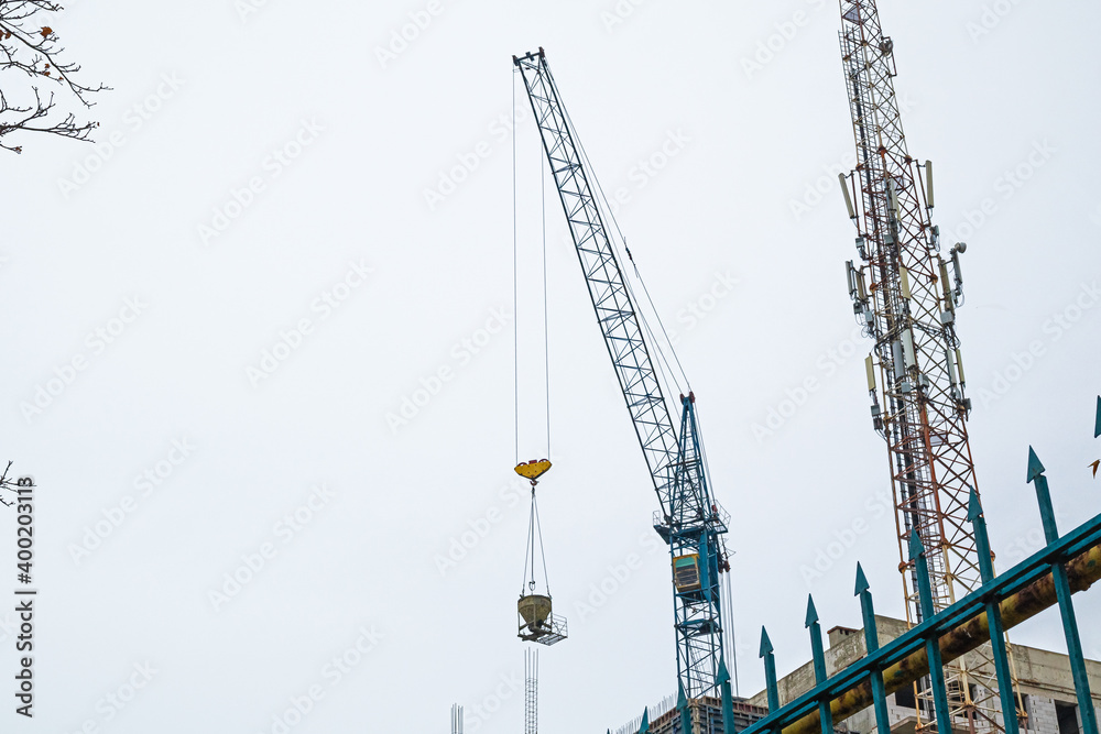 The Hoisting Tower Crane Moves the Concrete Mixer to The Workplace at The Construction Site. A Cell Phone Mast Is Visible Next to The Building Under Construction The Hoisting Tower Crane Moves the Concrete Mixer to The Workplace at The Construction Site. A Cell Phone Mast Is Visible Next to The Building Under Construction