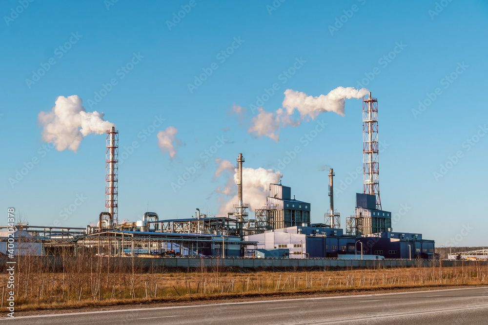 Pipes with huge clouds of smoke at the factory against the blue sky on a sunny day, photo from the road. Concept of ecology, environmental pollution