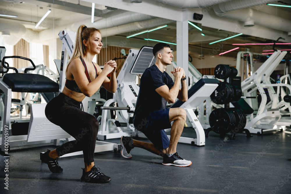 Fototapeta premium Two athletes perform squats. The coach shows the correctness of performing the exercises on the feet. Exercise in the gym. Joint training in small groups. A man and a girl train together.
