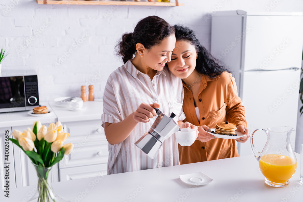 Hispanic same sex couple pouring coffee and holding pancakes near flowers on blurred foreground in kitchen Hispanic same sex couple pouring coffee and holding pancakes near flowers on blurred foreground in kitchen
