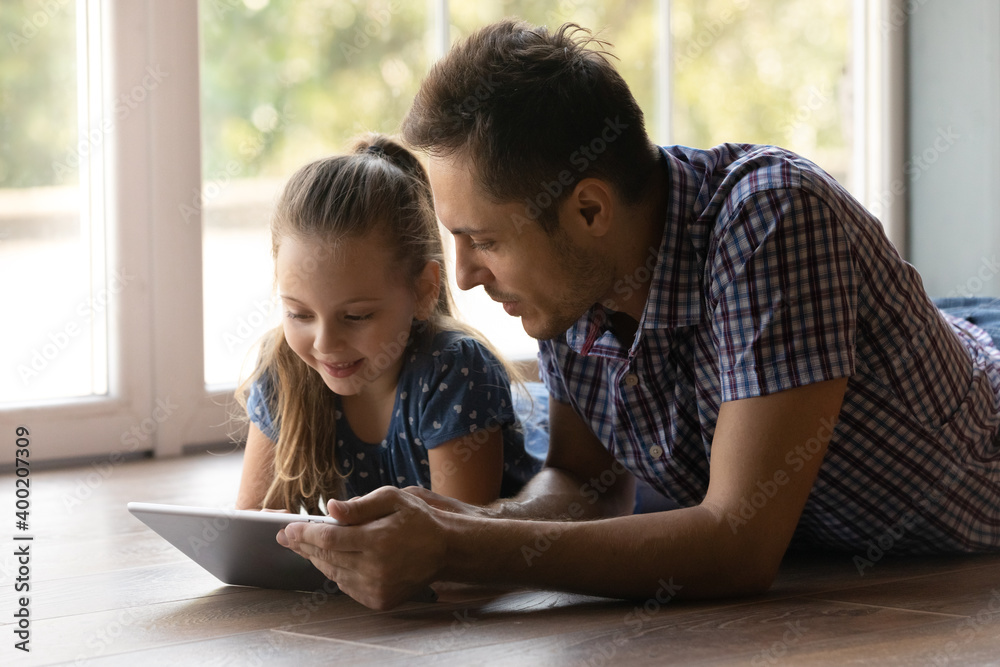 Shared hobby. Cute little daughter spend free time with young loving daddy lie on heated floor by window look at tablet screen. Junior schoolgirl read electronic book online browse web on pad with dad Shared hobby. Cute little daughter spend free time with young loving daddy lie on heated floor by window look at tablet screen. Junior schoolgirl read electronic book online browse web on pad with dad