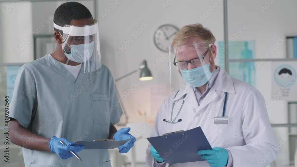 Portrait of Caucasian male doctor and his Afro-American assistant in medical uniform, protective mask, screen and gloves posing for camera in clinic while working during coronavirus outbreak Portrait of Caucasian male doctor and his Afro-American assistant in medical uniform, protective mask, screen and gloves posing for camera in clinic while working during coronavirus outbreak