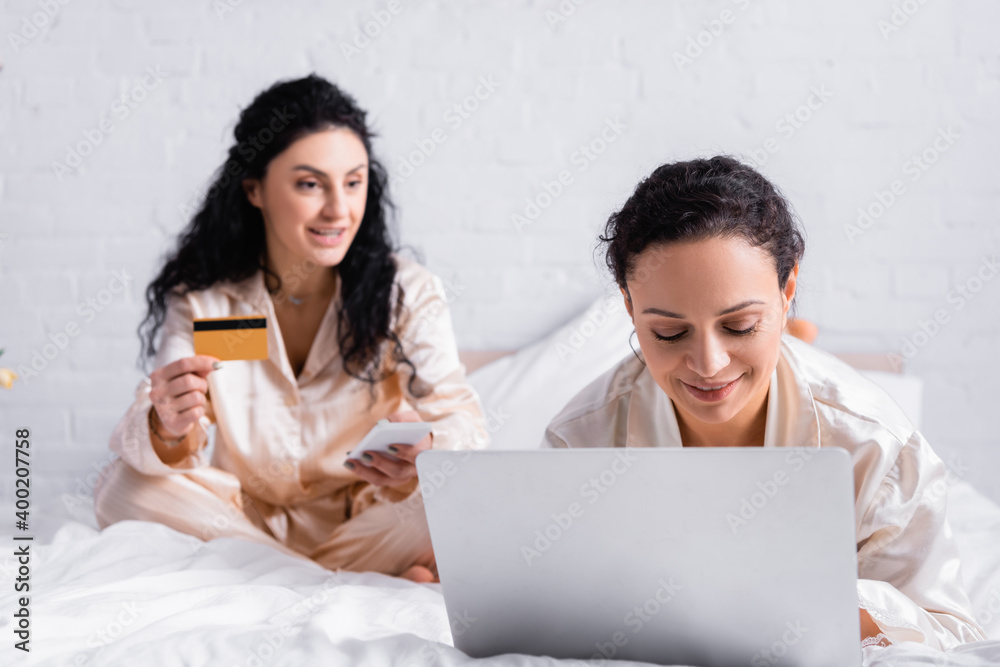 Hispanic woman using laptop near lesbian partner with credit card and smartphone on blurred background on bed Hispanic woman using laptop near lesbian partner with credit card and smartphone on blurred background on bed