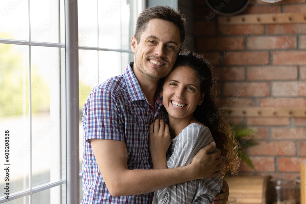 Cozy happiness. Family portrait of married millennial couple embrace with love care at new home house. Happy husband wife looking at camera glad receiving mortgage loan becoming homeowners first time Cozy happiness. Family portrait of married millennial couple embrace with love care at new home house. Happy husband wife looking at camera glad receiving mortgage loan becoming homeowners first time