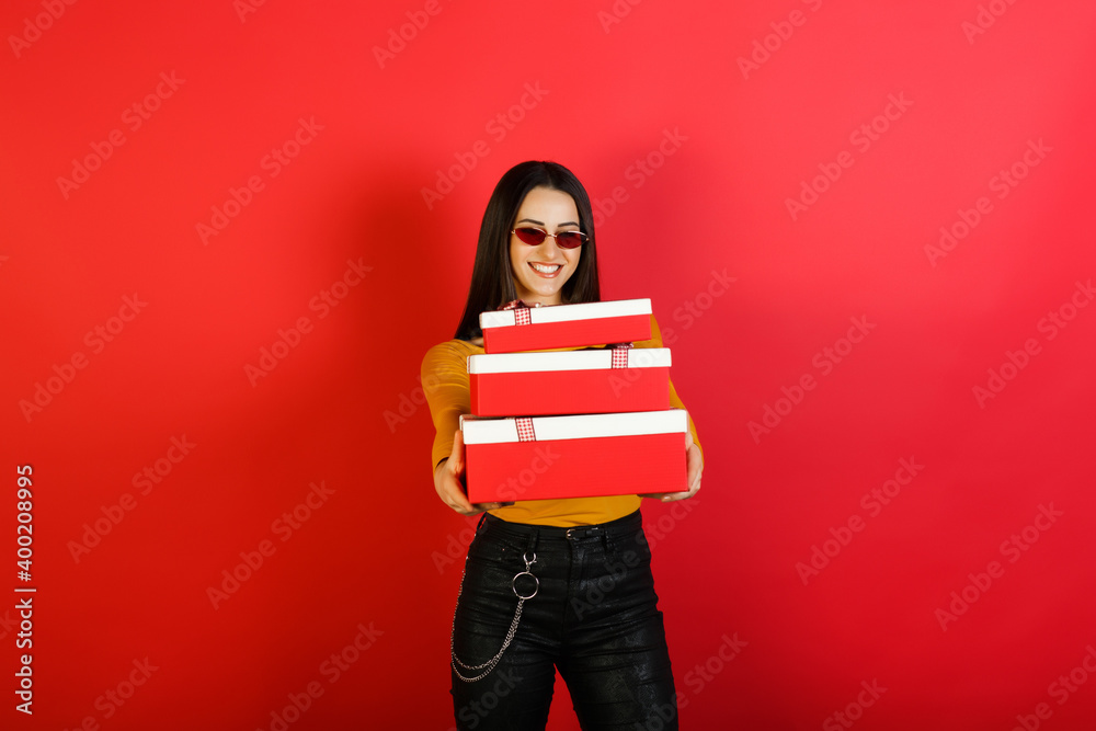 Young happy smiling woman in red sunglasses hold three red gift boxes isolated on red studio background. Young happy smiling woman in red sunglasses hold three red gift boxes isolated on red studio background.