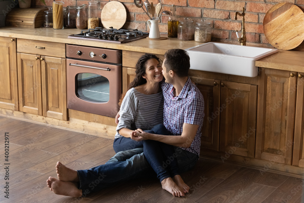 Tender loving young male husband sit barefoot on cozy wooden warm kitchen floor embrace cheering female wife hold her on knees look in eyes. Happy lovers enjoying date at home spending time together - obrazy, fototapety, plakaty 
