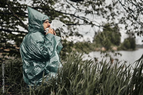 A man with a beard in a green raincoat eats in nature. Cloudy weather, fast food.