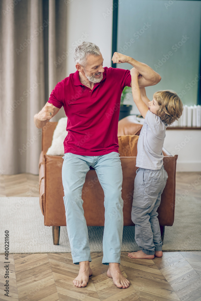 Grey-haired man showing his muscles to his son and feeling joyful