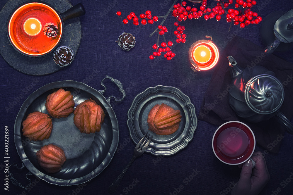 Obraz premium Yummy eclairs or brewing cakes and pewter tea pot, hand with tea cup. Christmas decor, candles with red berries. Xmas flat lay with sweets, tea and candles on dark purple background.
