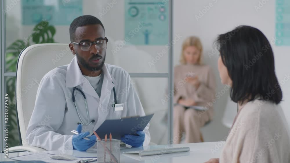 Young Afro-American male doctor in lab coat and gloves checking medical history and talking with female patient during consultation in clinic Young Afro-American male doctor in lab coat and gloves checking medical history and talking with female patient during consultation in clinic