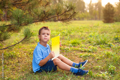 Wallpaper Mural Little boy a schoolboy in a park at sunset reads a book. A surprised joyful child in a blue shirt with a yellow book in the park sits on the grass at sunset. Distance learning has ended. Torontodigital.ca