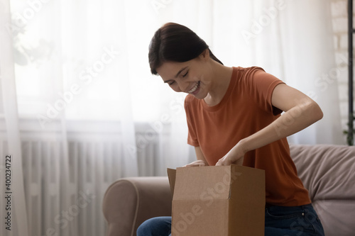 Pleasant surprise delivered by mail. Curious young woman shopper unpacking cardboard box getting order from online store. Happy excited millennial lady opening postal parcel amazed with gift received