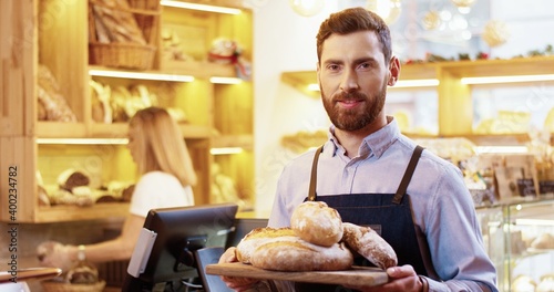 Close up of happy bearded young Caucasian man seller in apron standing in bakery and holding tray with fresh baked bread in hands, smelling baking and looking at camera while colleague works behind