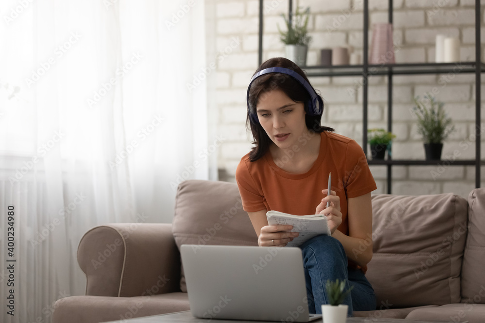 Attentive young woman sitting on couch by laptop listening to learning material in headphones. Female student study from home online at quarantine period taking notes of lecture to paper exercise book Attentive young woman sitting on couch by laptop listening to learning material in headphones. Female student study from home online at quarantine period taking notes of lecture to paper exercise book