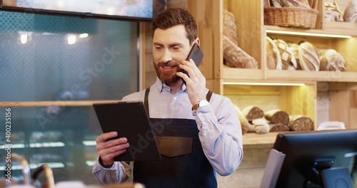 Photos Portrait of happy Caucasian bearded handsome male baker standing in own bakery, tapping on tablet device and speaking on smartphone