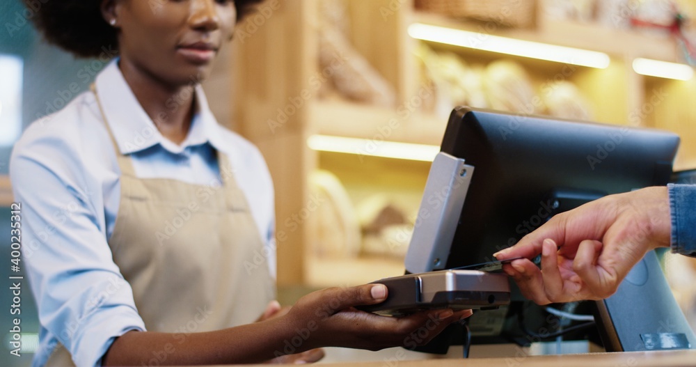 Close up of African American female seller in mask and protective gloves selling food in bakery shop. Client hand paying with credit card on device. Small business. Shopping. Bakehouse concept Close up of African American female seller in mask and protective gloves selling food in bakery shop. Client hand paying with credit card on device. Small business. Shopping. Bakehouse concept