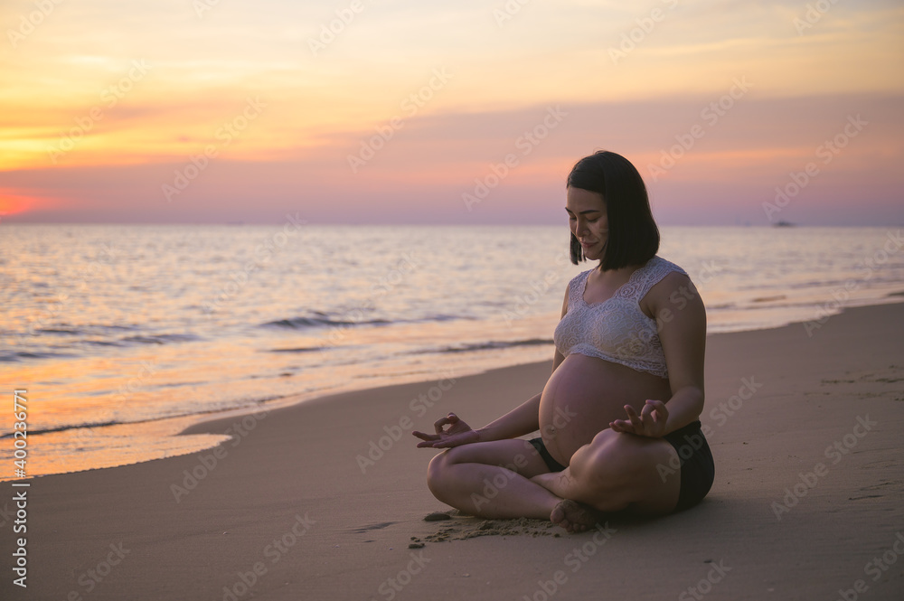 Young pregnant Asian woman doing Yoga on the beach at sunset.