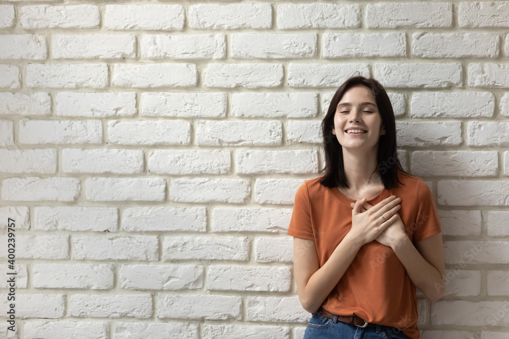 What a relief. Happy young woman posing close to wall of bricks with closed eyes feeling grateful hopeful. Millennial female putting hands on chest over heart express sincere feelings. Copy space