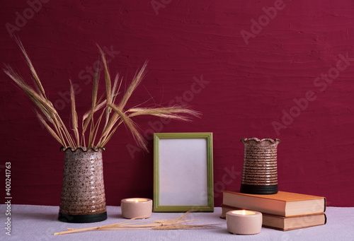 old books, candles are burning, vases with ears of wheat, dry daffodils, photo frame on a red background