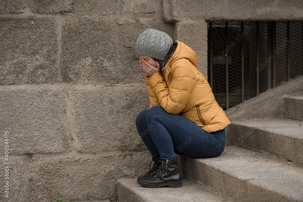 dramatic lifestyle portrait of young attractive sad and depressed Chinese woman in winter hat sitting outdoors on street corner staircase suffering depression problem crying
