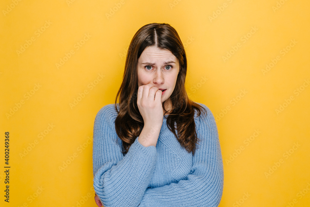 Close up portrait of nervous young caucasian woman biting her nails while looking at camera, dressed in blue knitted sweater,  isolated over yellow studio background. People emotion concept