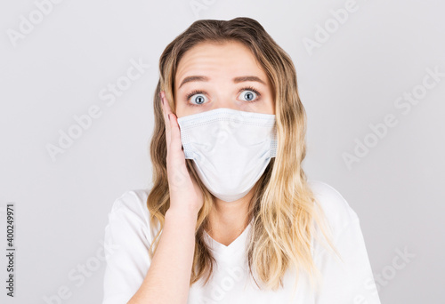 Portrait of a young surprised woman in a medical mask isolated over grey background. Young girl patient stands against the wall background wirh fear in her eyes.