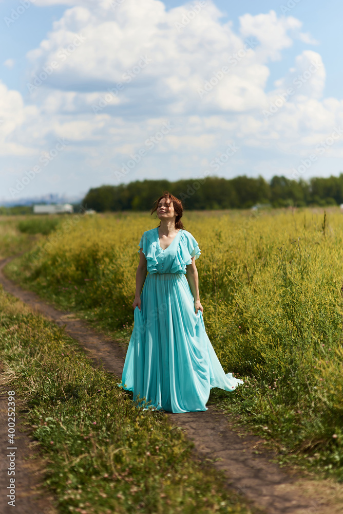 Young woman walking flowering field, turned away, gently touch yellow flowers. Brunette girl enjoys spring aromas, harmony. Backdrop dark clouds blue sky. Long hair fluttering wind. Denim sundress hat Young woman walking flowering field, turned away, gently touch yellow flowers. Brunette girl enjoys spring aromas, harmony. Backdrop dark clouds blue sky. Long hair fluttering wind. Denim sundress hat