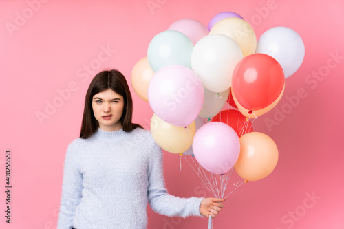 Young Ukrainian teenager girl holding lots of balloons over isolated pink background having doubts and with confuse face expression