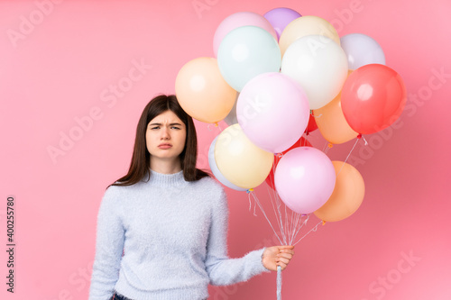 Young Ukrainian teenager girl holding lots of balloons over isolated pink background sad