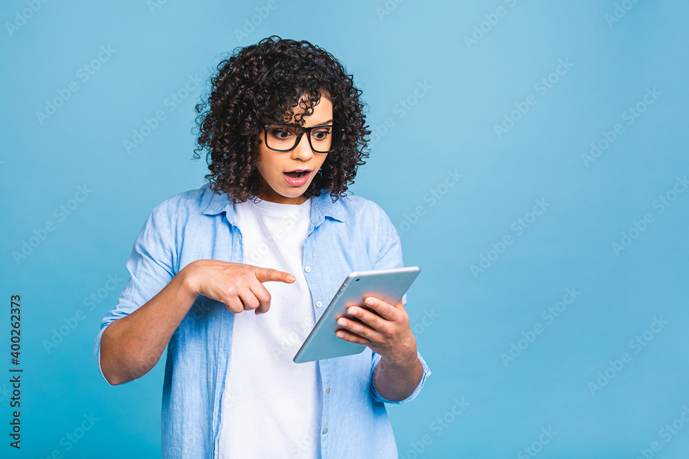 Shocked american student girl with curly african hair holding digital tablet over isolated blue background with copy space for text, logo or advertising. Shocked american student girl with curly african hair holding digital tablet over isolated blue background with copy space for text, logo or advertising.