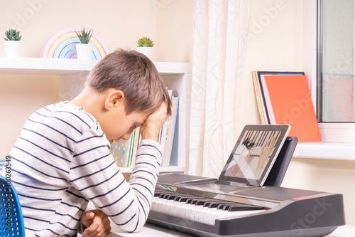 Sad frustrated boy has difficulty learning to play piano at home. Kid using digital tablet to watch video lesson. Online learning difficulties photo