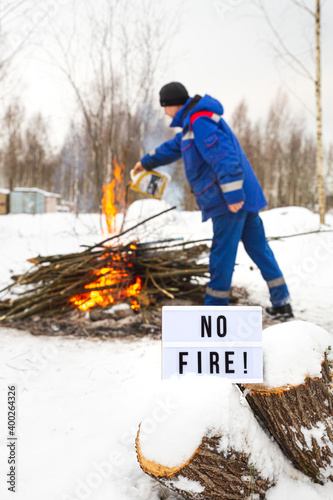 A man in a blue uniform pours gasoline from a canister into the fire. The concept of nature protection, protection from forest fires, ecology. Open fire warning. A sign with the text No fire.