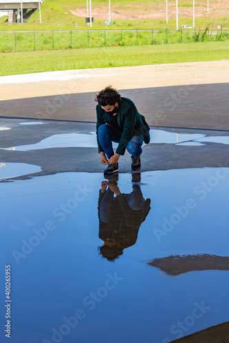 Homem elegante, usando máscara de proteção, abaixado, amarrando o sapato com reflexo em poça de água no chão. © Angela