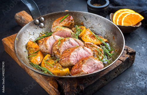 Traditional fried pork filet medaillons in with caramelized orange slices and herbs served as close-up in a rustic wrought iron skillet