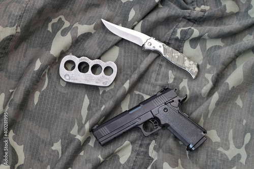 Handgun lies with brass knuckles and knife on camouflage military uniform close up. Concept of looting and arms trade photo