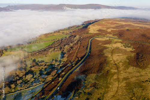 Aerial view of a narrow, winding mountain road emerging through a bank of fog and low cloud on a cold, frosty day
