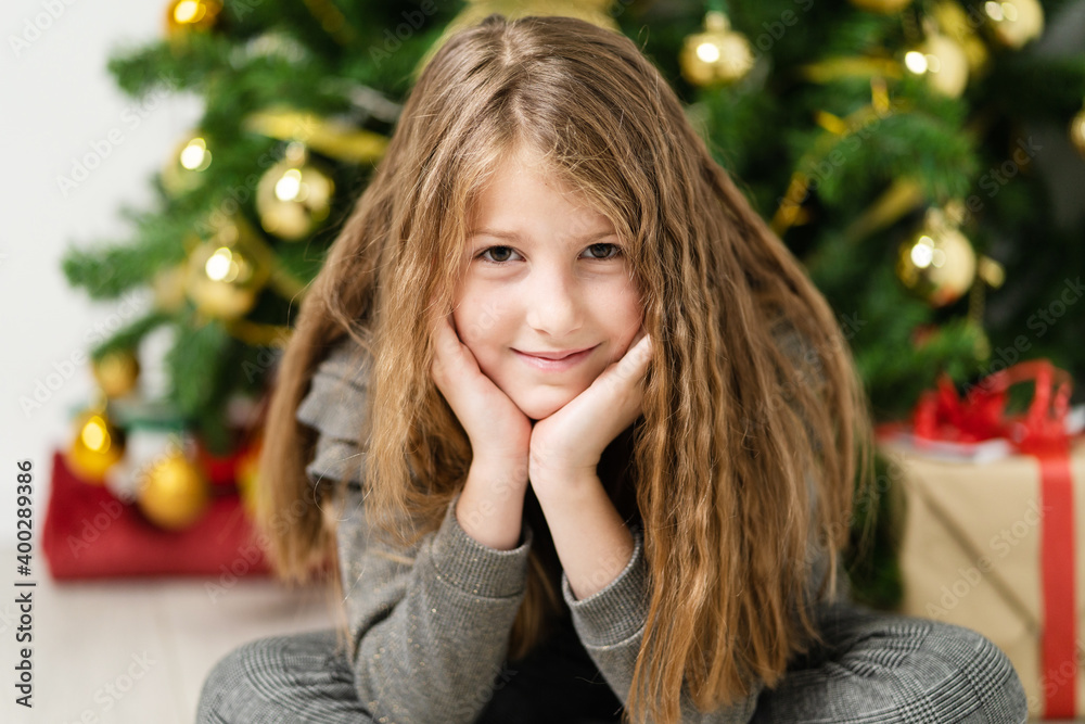 Front view portrait of small caucasian girl eight years old sitting in front of the christmas tree at home with wrapped box gift presents looking to the camera joyful Front view portrait of small caucasian girl eight years old sitting in front of the christmas tree at home with wrapped box gift presents looking to the camera joyful