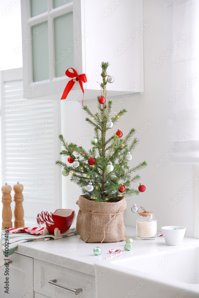 Fototapeta premium Small Christmas tree decorated with baubles and festive lights in kitchen