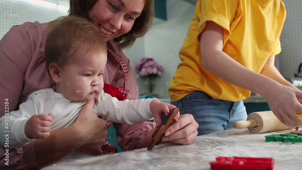 The mother holds the baby in her arms and shows her a gingerbread. Close-up portrait of a child. In the background, a woman rolls out the dough with a rolling pin. The concept of family cooking The mother holds the baby in her arms and shows her a gingerbread. Close-up portrait of a child. In the background, a woman rolls out the dough with a rolling pin. The concept of family cooking
