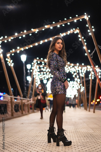 Christmas lifestyle. Brunette Caucasian girl in a fashionable sequined dress and black shoes with heels  in the city christmas lights
