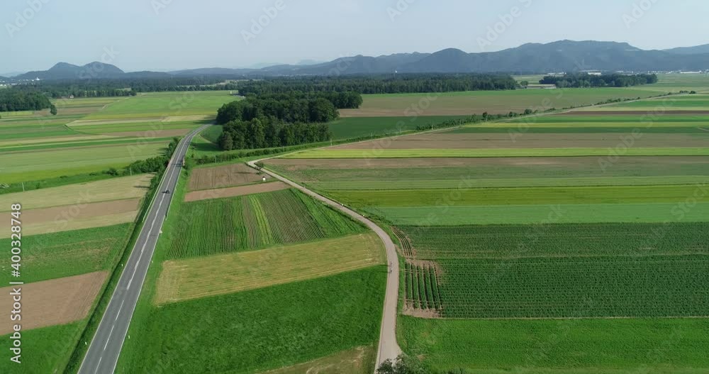Drone aerial view of green farming grassland field in Slovenia. Paved countryside road between agriculture fields. Ljubljana basin flatland with mountains in the distance. Descending Drone aerial view of green farming grassland field in Slovenia. Paved countryside road between agriculture fields. Ljubljana basin flatland with mountains in the distance. Descending