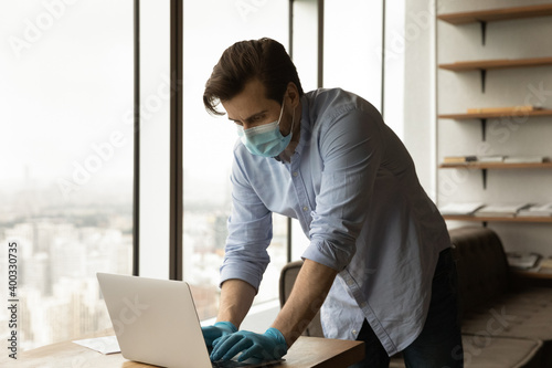 Young Caucasian male worker in medical facial mask and rubber gloves work on laptop in office during coronavirus pandemics. Man employee in protective gear use computer. Covid-19, corona concept. © fizkes