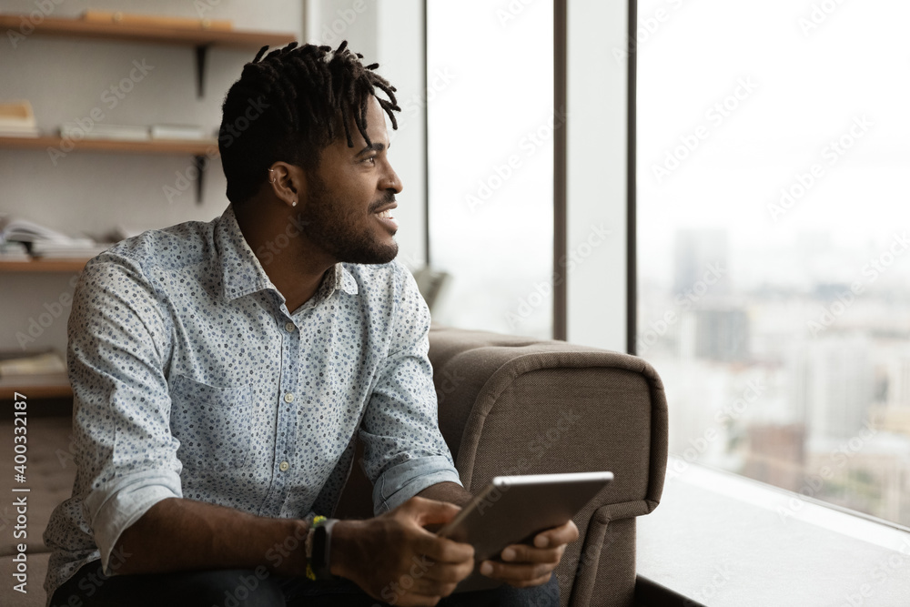 Smiling biracial man relax at home use modern tablet look in distance thinking or dreaming or perspectives. Happy African American male hold pad device make decision or plan. Technology concept. Smiling biracial man relax at home use modern tablet look in distance thinking or dreaming or perspectives. Happy African American male hold pad device make decision or plan. Technology concept.