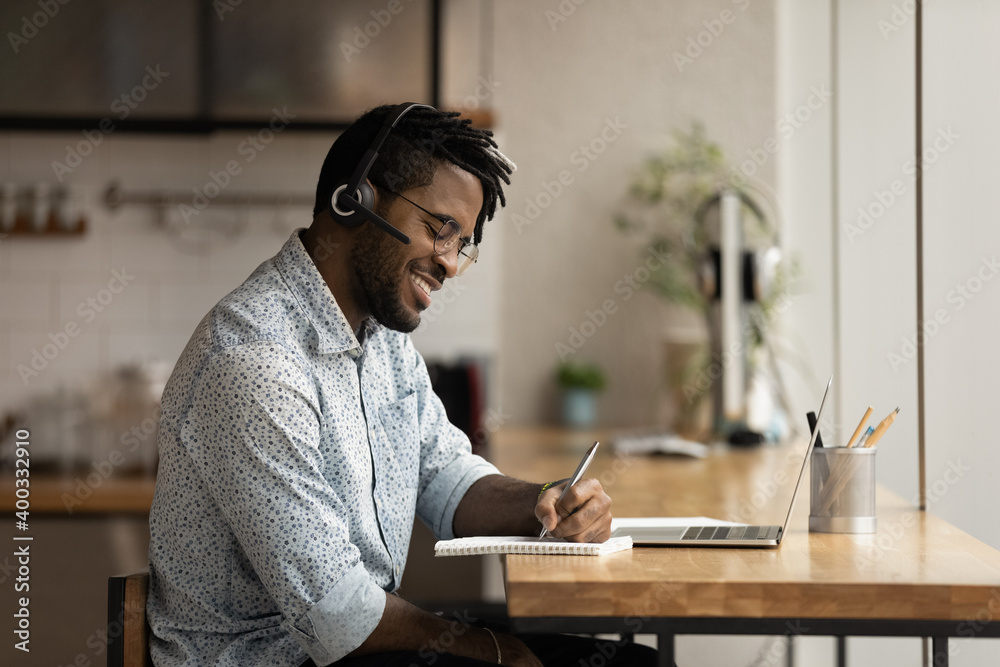 Fototapeta premium Smiling millennial African American man in headphones watch online webinar on laptop making notes. Happy young biracial male in earphones take distant course or study on computer. Education concept.