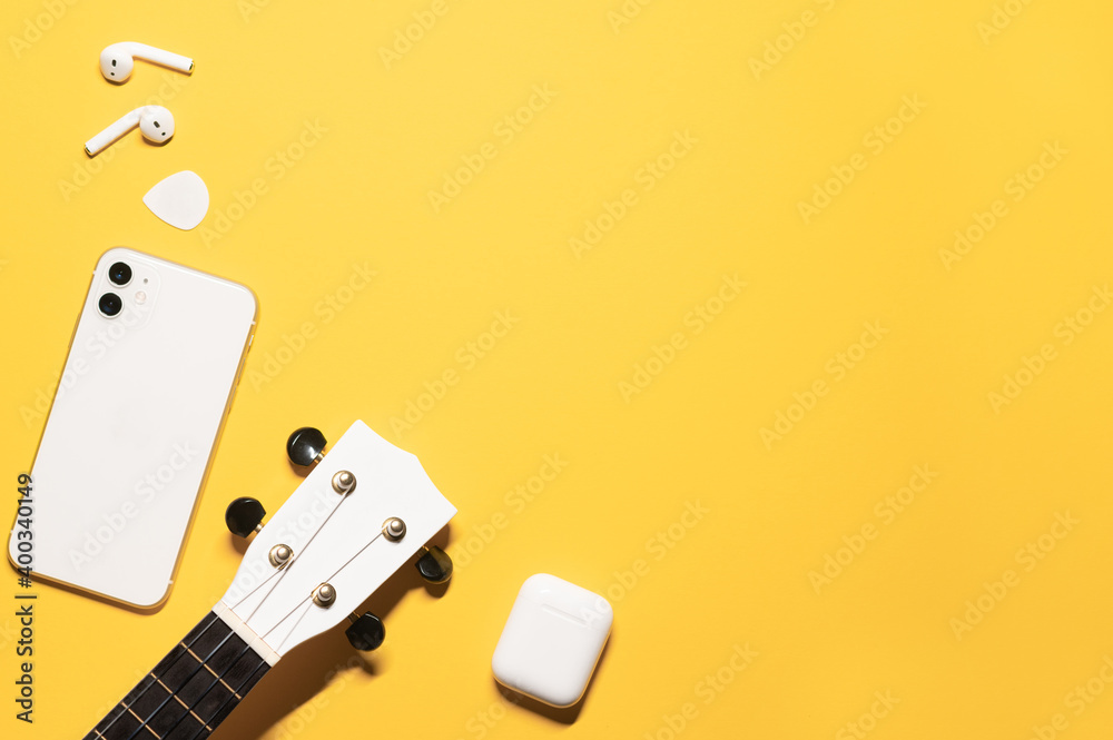 Flat lay composition with a copy space on a yellow background with a white ukulele fretboard, modern mobile phone, earphones with a charging case, and a guitar pick. Flat lay composition with a copy space on a yellow background with a white ukulele fretboard, modern mobile phone, earphones with a charging case, and a guitar pick.
